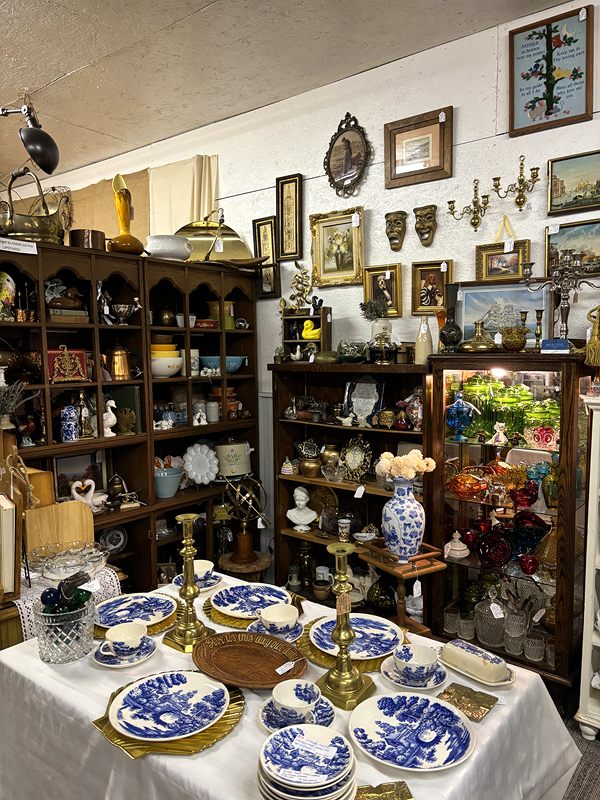 Blue-and-white plates and brass candlesticks on a table, with shelves of china and framed art behind