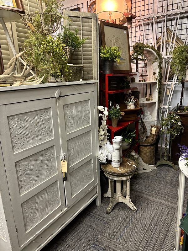 A white painted cabinet topped with greenery, beside a small pedestal table and garden décor