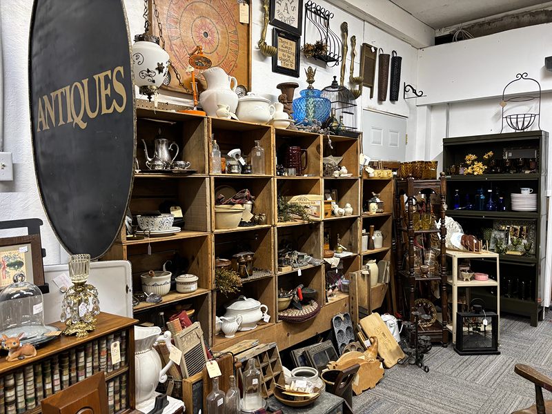 Cubby shelves of white pitchers, teapots and china beside a large round Antiques sign