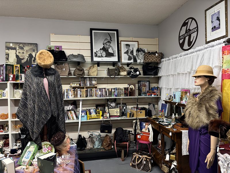 Mannequins in a fur stole and a cape, among purses, hats and framed photos